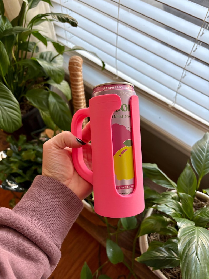 May include: A bright pink can cooler with a handle, holding a can of raspberry lemon sparkling drink. The can is visible through a cutout in the cooler. The cooler is held in front of a background of green plants.