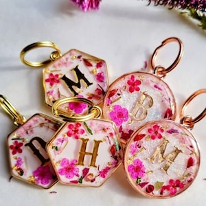 May include: Five keyrings with gold rings and hexagonal or round resin charms. The charms are filled with pink and white dried flowers and gold glitter. Each charm has a gold letter, M, B, H, and M.