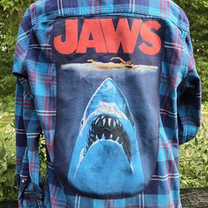 May include: Blue plaid flannel shirt with a large graphic of the movie "Jaws" on the back. The graphic features the word "JAWS" in red letters above an image of a shark and a swimmer.