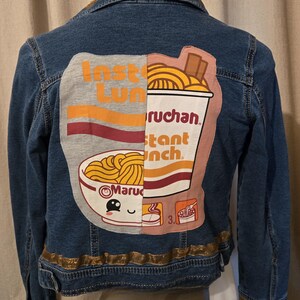 May include: Blue denim jacket with a large graphic on the back featuring a split design of instant ramen noodles. The graphic includes the words "Insta Lun" and "Instant Lunch" with a bowl of noodles and a smiling face.