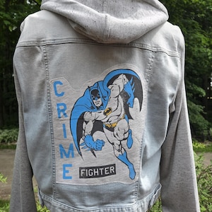 May include: A light gray denim jacket with a gray hood. The back of the jacket features a large graphic of Batman in blue, black, and yellow, with the words "CRIME FIGHTER" in blue.
