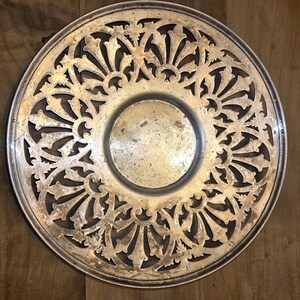 May include: A silver-toned, round trivet with an intricate openwork design. The trivet features a central, slightly recessed area and a decorative pattern of stylized floral or leaf-like shapes radiating outwards. The metal has a slightly aged appearance.