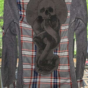 May include: Gray flannel shirt with a plaid pattern in red, white, and blue. The back features a large, dark gray graphic of a skull and snake design. The sleeves are a solid gray material.