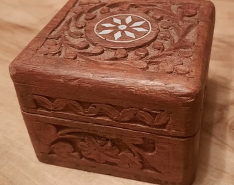 May include: A small, intricately carved wooden box with a floral design. The box has a white inlay in the center of the lid, featuring a starburst pattern.