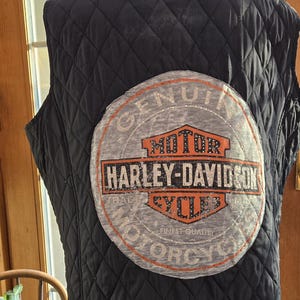 May include: Black quilted vest with a large Harley-Davidson patch on the back. The patch is a circular design with the Harley-Davidson logo in orange and white. The vest has a diamond pattern.