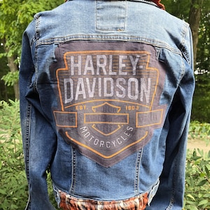 May include: Blue denim jacket with a Harley-Davidson logo on the back. The jacket features a plaid fabric trim and collar. The Harley-Davidson logo is in orange and gray. The jacket is displayed on a mannequin.