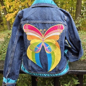 May include: A denim jacket with a large, colorful butterfly design on the back. The butterfly features pink, orange, yellow, green, blue, and purple hues with gold accents. The jacket has a teal collar and trim.