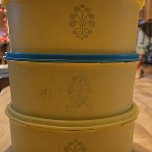 May include: Three yellow plastic containers with blue lids stacked on top of each other. Each container has a decorative design of a stylized tree with a circle at the top.