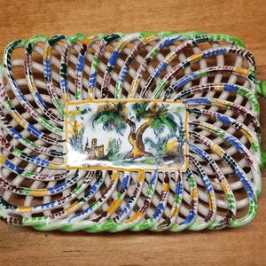May include: A white ceramic serving dish with green, blue, and yellow painted details. The dish has a woven design and two green handles. A painted scene of a tree and a house is in the center of the dish.