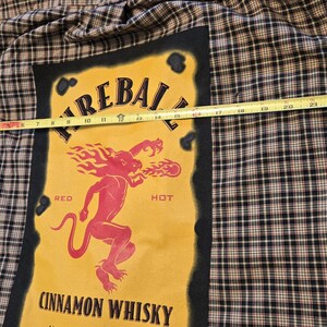 May include: A plaid button-up shirt with a large yellow graphic featuring the text "FIREBALL CINNAMON WHISKY". The graphic includes a red devil-like figure breathing fire. The shirt is approximately 23 inches wide.