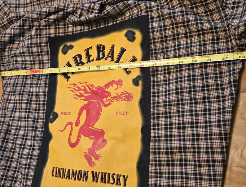 May include: A plaid button-up shirt with a large yellow graphic featuring the text "FIREBALL CINNAMON WHISKY". The graphic includes a red devil-like figure breathing fire. The shirt is approximately 23 inches wide.