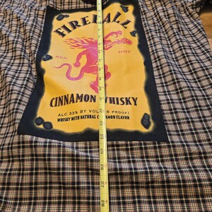 May include: A plaid flannel shirt with a large graphic featuring the text "Fireball Cinnamon Whisky." The graphic is yellow with a red and pink illustration. The shirt is approximately 30 inches long.