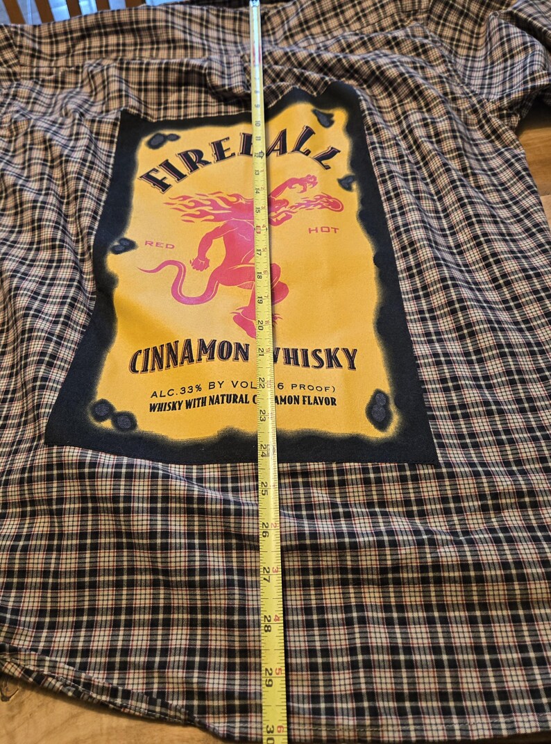 May include: A plaid flannel shirt with a large graphic featuring the text "Fireball Cinnamon Whisky." The graphic is yellow with a red and pink illustration. The shirt is approximately 30 inches long.