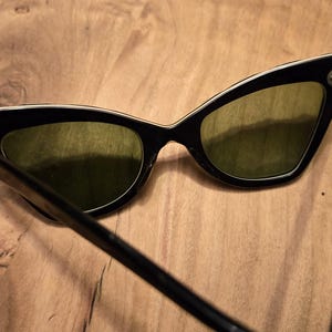 May include: Black cat-eye sunglasses with green lenses and a white trim around the edges. The sunglasses are on a wooden surface.