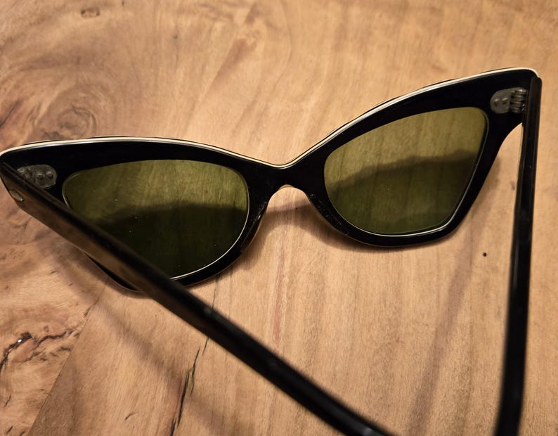 May include: Black cat-eye sunglasses with green lenses and a white trim around the edges. The sunglasses are on a wooden surface.