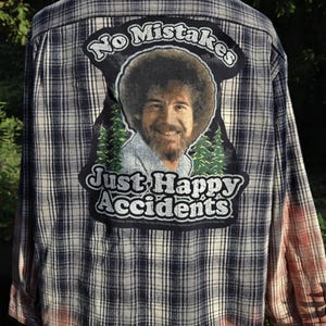 May include: Plaid button-down shirt with a faded ombre effect at the bottom. The back features a patch with Bob Ross's image and the text "No Mistakes, Just Happy Accidents."