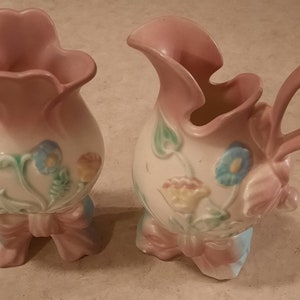 May include: Two vintage ceramic vases with floral designs and pink and white glaze. The vases have a ruffled top and a bow at the base.