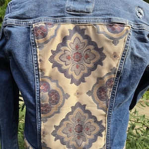 May include: A denim jacket with a decorative back panel. The back panel features a cream-colored fabric with a repeating floral pattern in shades of blue, red, and gold. The jacket has a classic collar and button closures.