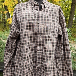 May include: A button-down plaid shirt in a brown, black, and white pattern. The long-sleeved shirt has a button-down collar and a single chest pocket. The shirt is displayed on a mannequin against a backdrop of fall foliage.