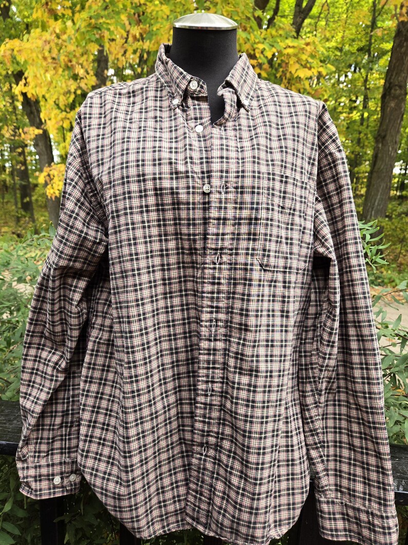 May include: A button-down plaid shirt in a brown, black, and white pattern. The long-sleeved shirt has a button-down collar and a single chest pocket. The shirt is displayed on a mannequin against a backdrop of fall foliage.