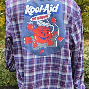 May include: A purple, blue, and white plaid flannel shirt with a large patch featuring the Kool-Aid Man. The patch includes the words "Kool-Aid" and "Oh, Yeahhh!" with an image of the Kool-Aid Man holding a pitcher.