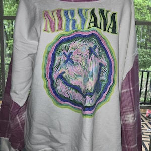 May include: White sweatshirt featuring the word "NIRVANA" in colorful letters above a smiley face design. The sleeves are a pink and white plaid pattern. The neckline is also trimmed with the plaid fabric.