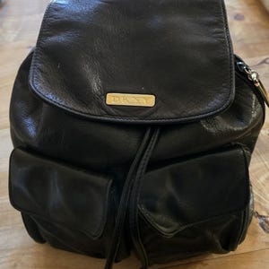 May include: Black leather backpack with a flap closure and two front pockets. The backpack features a drawstring closure and a small gold-toned metal plate with the letters "DKNY". The bag has a top handle.