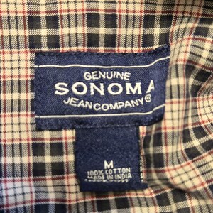 May include: Close-up of a plaid shirt with a navy blue label. The label reads "GENUINE SONOMA JEAN COMPANY" and "M 100% COTTON MADE IN INDIA". The shirt has a pattern of navy blue, white, and burgundy squares.