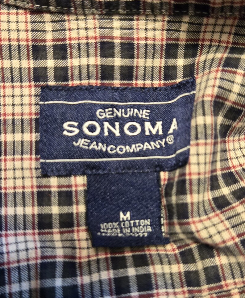 May include: Close-up of a plaid shirt with a navy blue label. The label reads "GENUINE SONOMA JEAN COMPANY" and "M 100% COTTON MADE IN INDIA". The shirt has a pattern of navy blue, white, and burgundy squares.