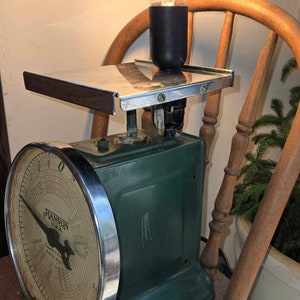 May include: Vintage green Hanson postal scale repurposed as a lamp. The scale features a silver-colored tray and a large circular dial with black numerals. A black lamp holder supports a decorative Edison-style light bulb. The lamp sits on a wooden chair.