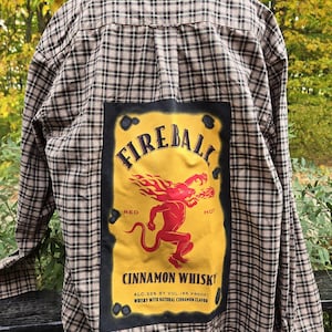 May include: Plaid button-down shirt with a large Fireball Cinnamon Whisky patch on the back. The patch features a red devil-like figure with flames, the brand name, and text indicating the alcohol content and flavor.
