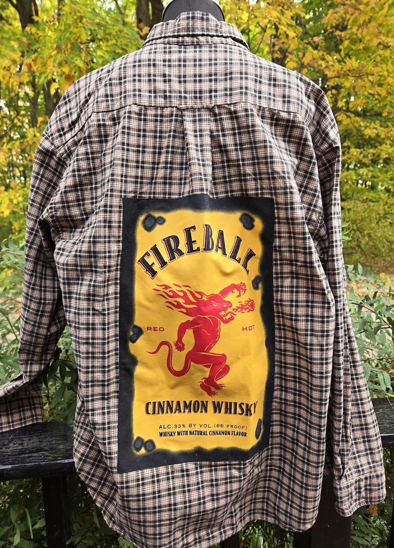 May include: Plaid button-down shirt with a large Fireball Cinnamon Whisky patch on the back. The patch features a red devil-like figure with flames, the brand name, and text indicating the alcohol content and flavor.