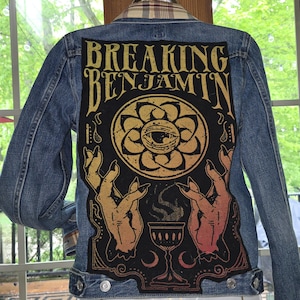 May include: A blue denim jacket with a large graphic on the back. The graphic features the words "BREAKING BENJAMIN" in gold lettering above a black and gold design with an eye, hands, and a chalice.