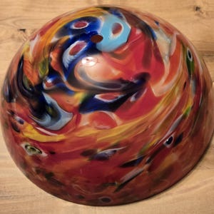 May include: A colorful, glass bowl with a swirling pattern of red, orange, yellow, blue, and white. The bowl has a rounded shape and a small hole at the top. The glass appears to be hand-blown.