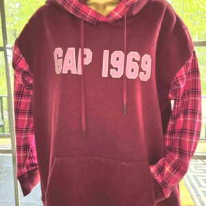 May include: A maroon hoodie with the text "GAP 1969" in pink. The sleeves and hood lining feature a pink and red plaid pattern. The hoodie has a front pocket and drawstrings.