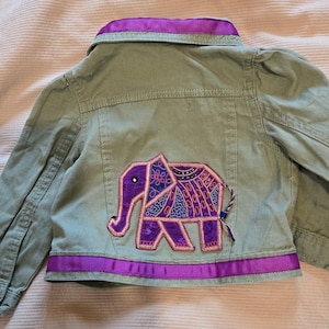 May include: Sage green denim jacket with purple trim and an embroidered elephant design on the back. The elephant is purple and blue with intricate patterns. The jacket has a button closure and long sleeves.