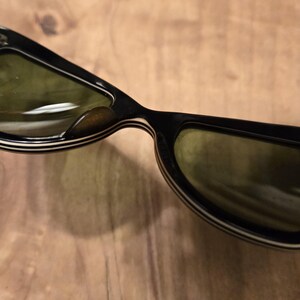 May include: Black sunglasses with white stripes on the frame and green lenses. The sunglasses are folded in half.
