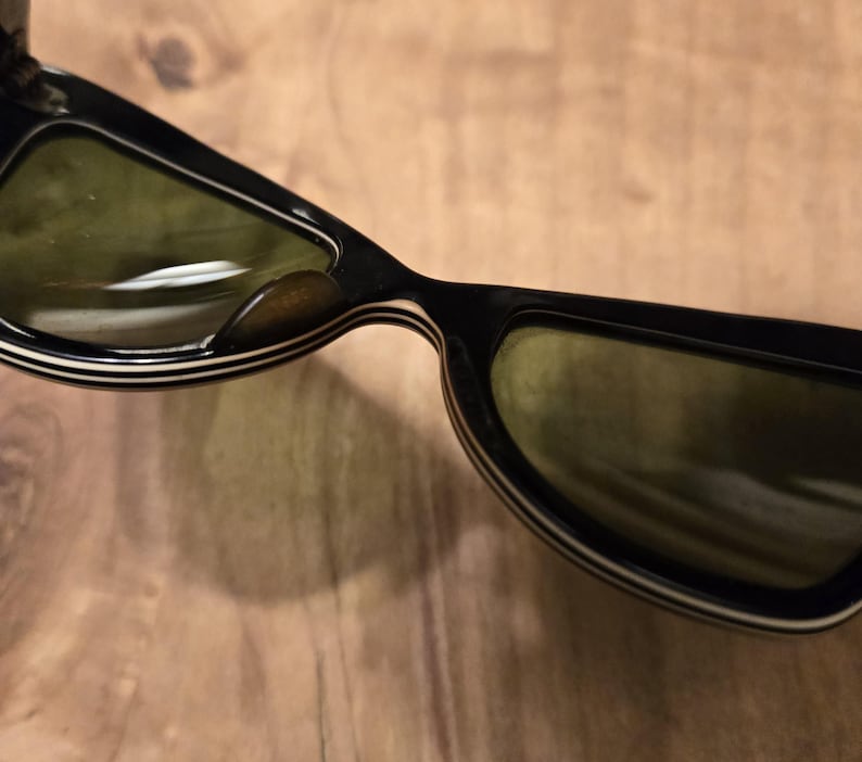 May include: Black sunglasses with white stripes on the frame and green lenses. The sunglasses are folded in half.