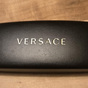 May include: A black rectangular glasses case with the gold-colored word "VERSACE" centered on the top. The case has a textured surface and a rounded shape.