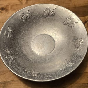 May include: A silver-colored decorative bowl with embossed grape and leaf designs. The bowl has a textured surface and a shallow, circular center. The grape and leaf designs are evenly spaced around the bowl's rim.
