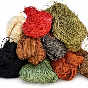 Puede incluir: Una colección de varios manojos de cordel fino y fibroso en diversos colores, incluyendo negro, crema, rojo brillante, verde oliva, marrón oscuro, gris y naranja terracota, apilados en un arreglo suelto y enredado sobre un fondo blanco.