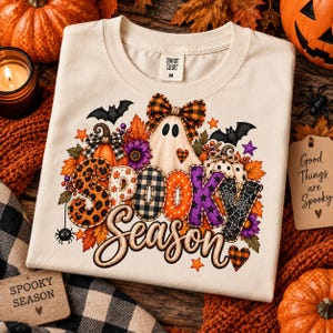 Könnte beinhalten: Ein cremefarbenes T-Shirt mit einem Grafikdruck und dem Text Spooky Season. Das Design zeigt ein Gespenst mit einer karierten Schleife, Kürbisse, Fledermäuse, Herbstblätter und Sterne. Die Buchstaben sind mit verschiedenen Mustern wie Leopardenprint gefüllt.