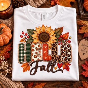 Könnte beinhalten: Ein weißes T-Shirt mit einer Grafik einer großen Sonnenblume, umgeben von Herbstblättern und Beeren. Der Schriftzug "HELLO Fall" ist in verschiedenen Mustern wie grünem Schottenkaro, Blumen, Rot mit Herzen und Leopardenmuster gestaltet, inklusive zwei Herzen.