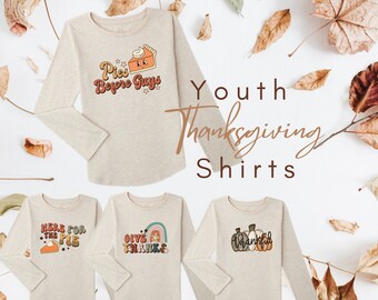 May include: Four long-sleeved shirts with Thanksgiving-themed designs. The shirts are beige with different graphics, including a pie, pumpkins, and a rainbow. The text on the shirts includes "Pies Before Guys", "Here for the Pie", "Give Thanks", and "Thankful".