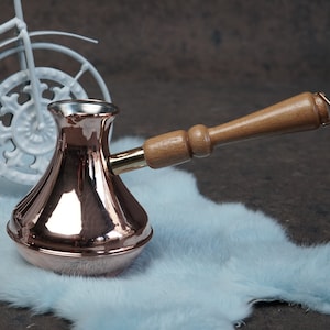 May include: A copper coffee pot with a wooden handle. The pot is sitting on a light blue fuzzy surface.