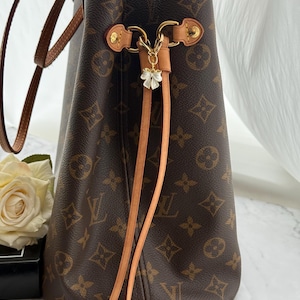 May include: Brown tote bag featuring a repeating monogram pattern. It has tan leather handles and trim, with gold-tone hardware. A small charm with a white flower hangs from the bag's closure. The interior lining is a bright pink colour.