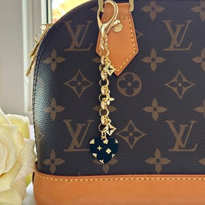 Puede incluir: Un charm de cadena de oro con un colgante de corazón negro con un patrón de monograma de Louis Vuitton. El charm está unido a un bolso de mano de cuero marrón con un cierre dorado.