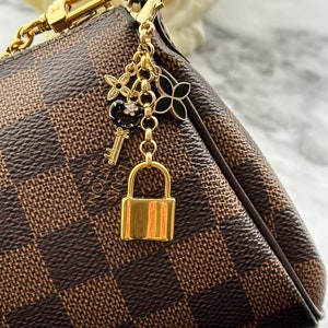 May include: A gold-tone handbag charm with a padlock, keys, and other decorative charms. The charm is attached to a brown and black checkered handbag.
