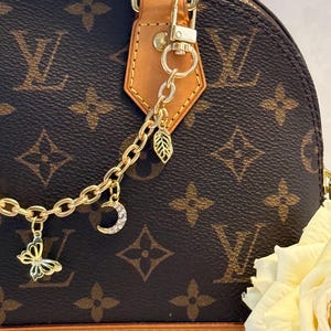 Può includere: Una catena di ciondoli color oro con ciondoli a forma di farfalla, mezzaluna e foglia attaccata a una borsa marrone e beige. La borsa ha un motivo ripetuto di loghi marroni su sfondo marrone.
