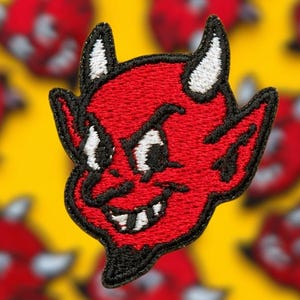 May include: An embroidered patch featuring a red devil character with white horns, pointed ears, and a black goatee. The design includes black outlines, white eyes, and a mischievous facial expression with a visible tooth, rendered in thread.